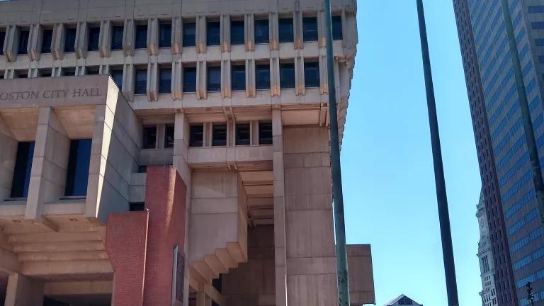 Photograph of young woman riding a cargo bike in front of Boston City Hall on a blue sky day with American and Boston City flags flying