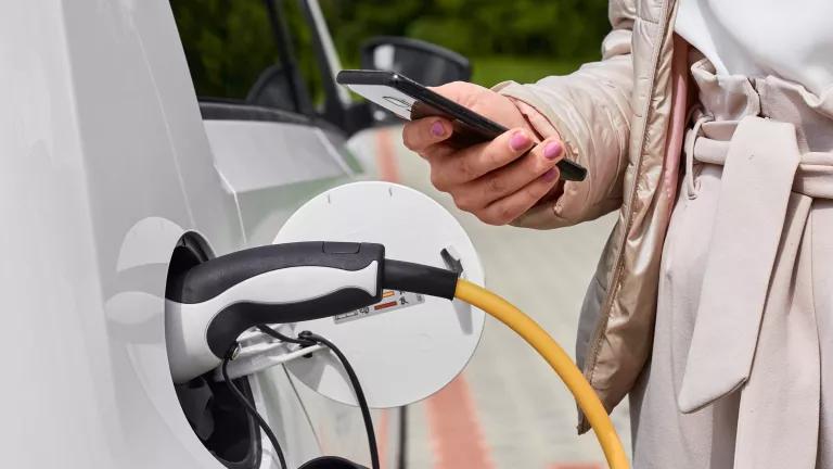 A person looks at a cell phone while standing next to a charging electric vehicle 