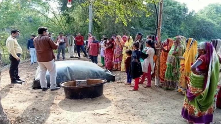 Villagers, primarily women and children in colorful clothing, stand by a biogas plant during a capacity building session 