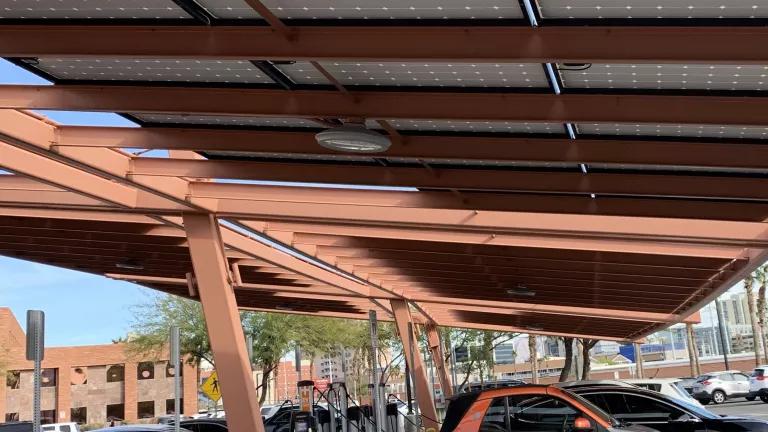 A small silver and orange electric car plugged in and charging in public parking lot in Las Vegas. The covering for the parking lot is made of solar panels.
