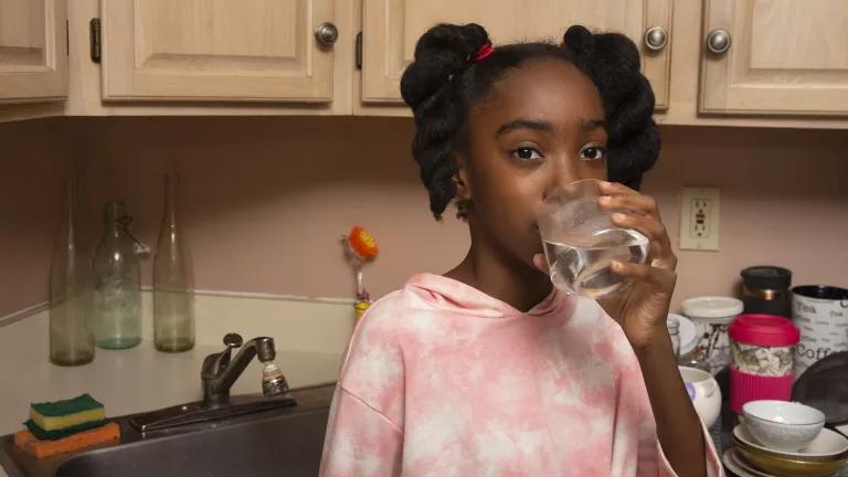 A Black pre-teen girl with braided pigtails and tie-dyed shirt is drinking a glass of tap water in front of a kitchen sink