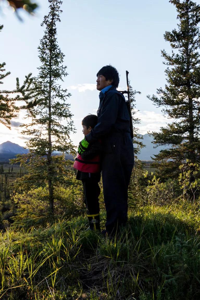 A man and young boy standing on a grassy hill overlooking an Alaskan landscape