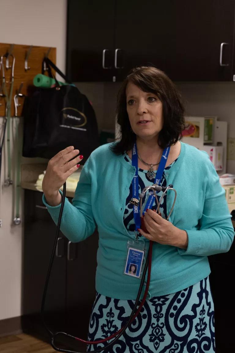 A person in a bright teal green cardigan and teal and black dress holding a stethoscope standing in front of a panel with hanging stethoscopes
