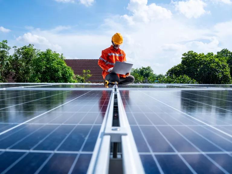 A person in an orange construction suit sitting on top of solar panels with a laptop