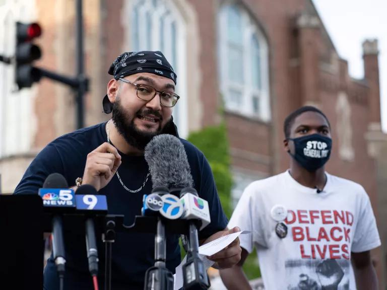 Oscar Sanchez speaks at an outdoor podium into microphones from several news channels with a young man behind him wearing a mask that reads “Stop General Iron” and a shirt reading, “Black Lives Matter.”