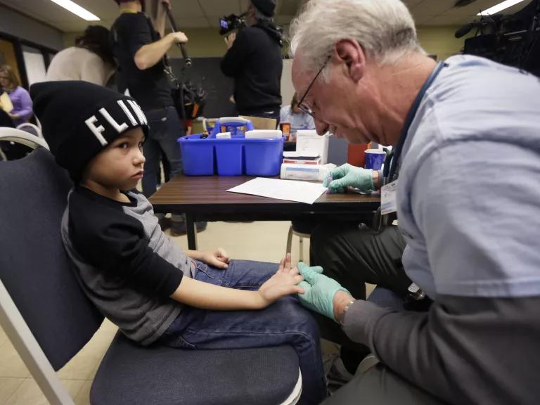 A young child sits in a chair having his finger pricked for a blood sample by a nurse in an elementary school.
