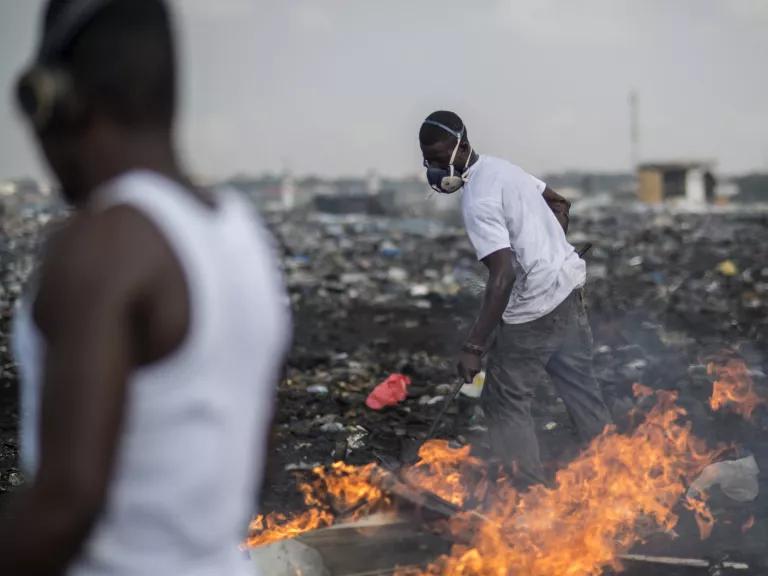 Young men on a large dumpsite shifting waste with an instrument as a fire burns the trash.