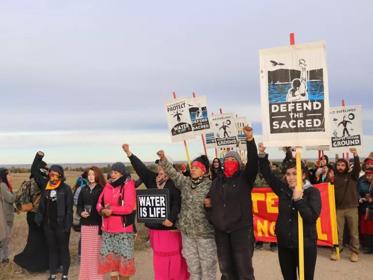 A group of protestors are marching outdoors along a dirt road holding signs that read, “Defend the Sacred” and “Water is Life.”