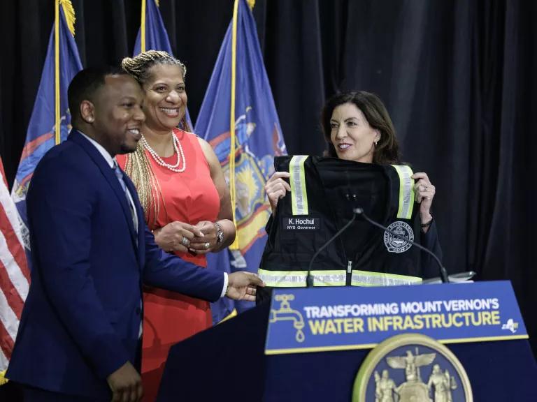 Government officials stand at a podium as one hands another a custom construction vest.