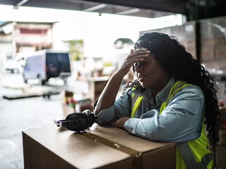 A worker in a warehouse leaning on a box and wiping her forehead.