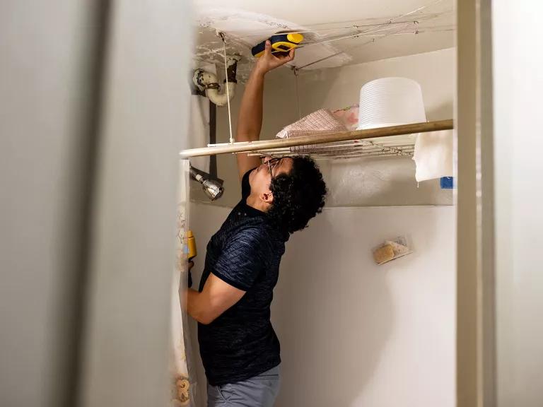 A man stands in a shower stall and holds a tool up to the ceiling where fresh spackle is visible