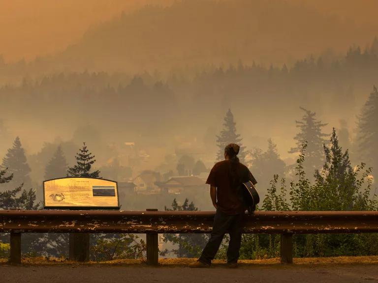 An Indigenous man looks out across the Columbia River as thick, orange-tinted wildfire smoke darkens the sky in Oregon.