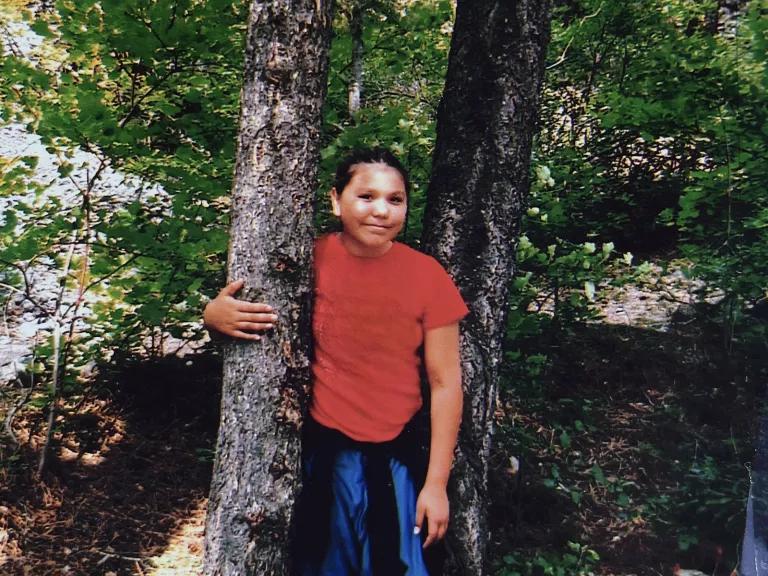A child in a red shirt and blue bottoms with one arm around a tree