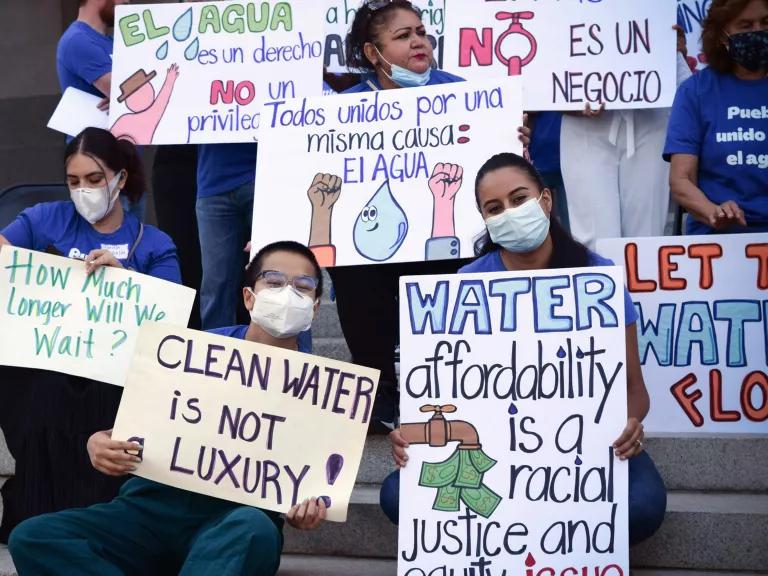 People on the steps of the state capitol in Sacramento hold signs saying “Clean water is not luxury!”; “Water affordability is a racial justice issue”; “Todos unidos por una misma causa: el auga”; and “How much longer will we wait?”
