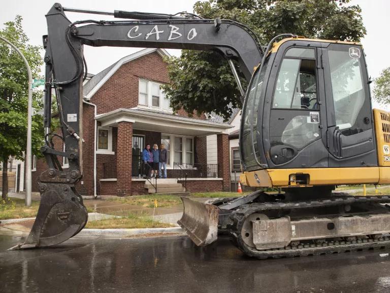 Gina Ramirez and her parents standing on the porch of their home, an excavator in the foreground