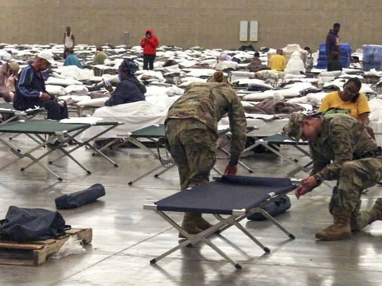 Two people in army fatigues set up rows of cots in a large space.