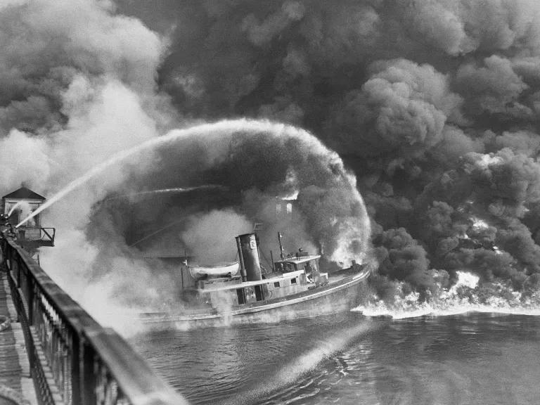 A black and white photo of firefighters hosing down a boat in a waterway