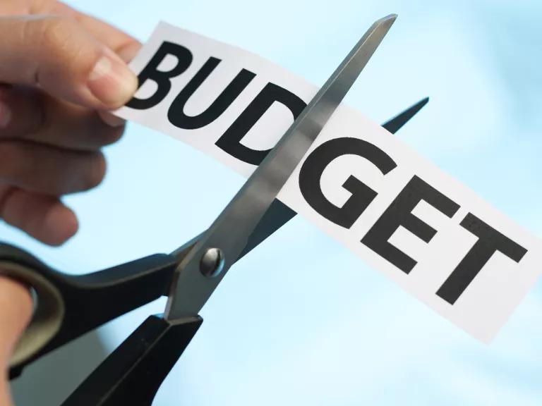 A person cutting a piece of paper with the word "budget" in half