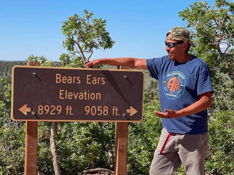 A person in a blue t-shirt and khaki pants wearing a cap and sunglasses pointing to a sign with their hand that says "Bears Ears Elevation"