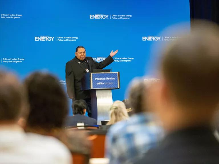 Wayne Yazza, Jr. stands on stage speaking to a crowd. He is standing behind a podium with his hand up, gesturing to something off screen.