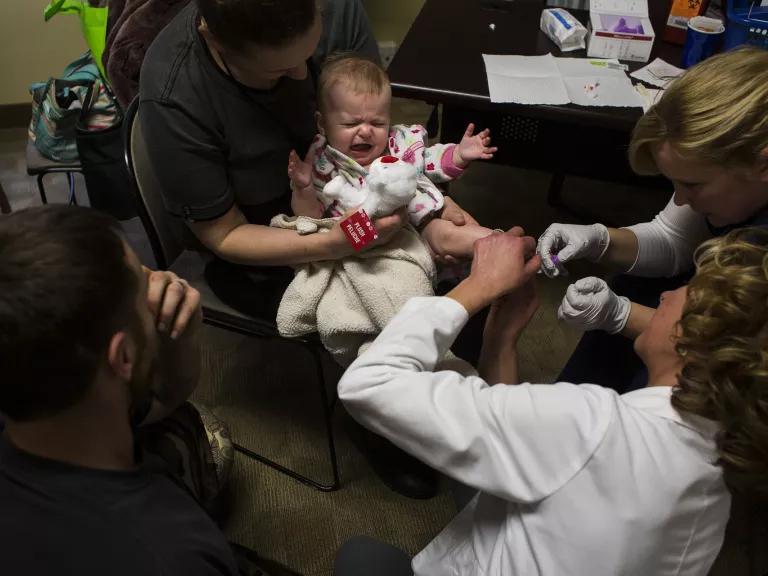 An adult holds a crying infant in her lap while two medical personnel admminister an injection in the infant's foot.
