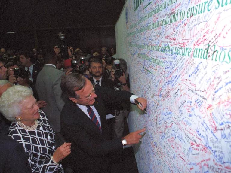 Former U.S. President George H.W. Bush signs a large white board covered in signatures, while Barbara Bush looks on.