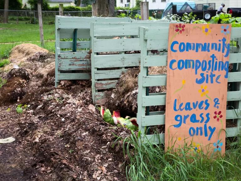 Open wooden bins in a grassy area with a painted sign that reads "Community composting site; Leaves & grass only"