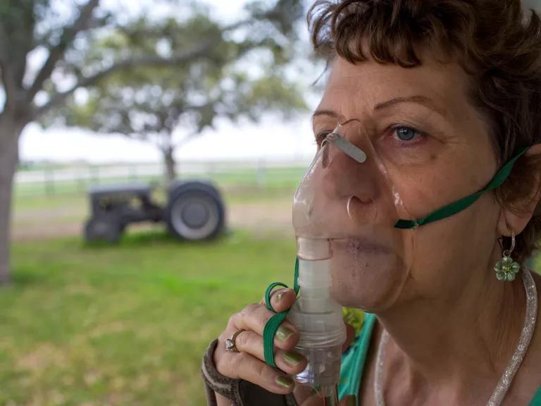 A woman holds an oxygen masks over her mouth and nose, with a view of a tree and tractor in a field behind her