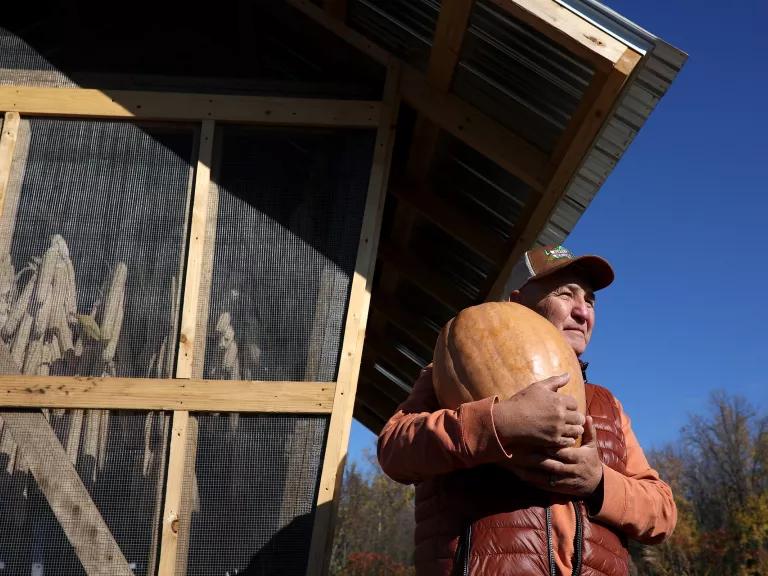 A man in a cap and outdoor vest in front of a wooden building holds a large squash