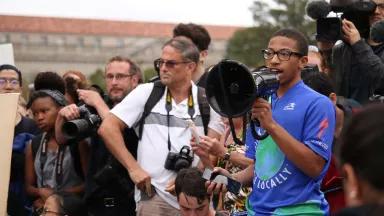 A young man speaks through a megaphone, with photographers behind him