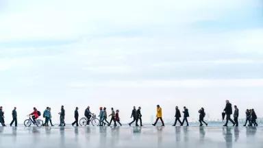 People walk on a paved walkway near a body of water with a city skyline in the background