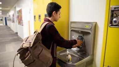 A junior high student filling up his water bottle at a drinking fountain in the school's hallway