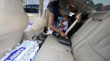 A Black woman leans over her daughter, who is drinking from a baby bottle, to buckle her seat belt. In the back seat of the car with the child is a case of bottled water.