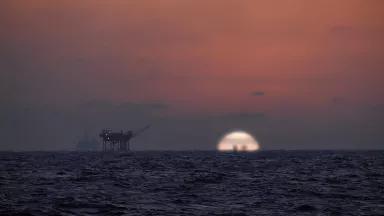 The sun is setting into the horizon of the Gulf of Mexico, with two offshore oil rigs are silhouetted by a red sky