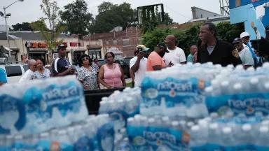 Cases of bottled water sit in the foreground with a line of people forming nearby.