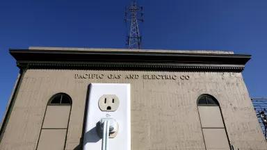 A low angle view of a truck tower carrying electrical lines behind an electrical substation with a sculpture of an electrical plug and socket on its wall