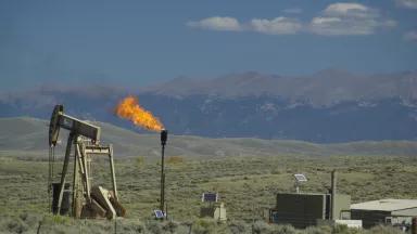 An oil well sits in a vast open space with low brush covering the ground and mountains in the distance