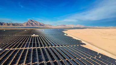 Dozens of long rows of solar panels are installed on a flat expanse of desert land with mountains in the background