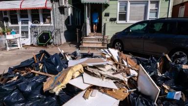 A large pile of debris and damaged building materials sits on the street in front of a house