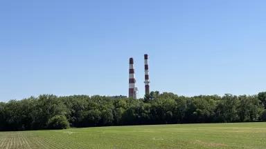 A green grassy fields with trees and smokestacks in the background against a blue sky