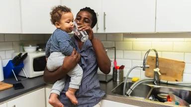 A mother gives her baby a sip of water from their kitchen sink.