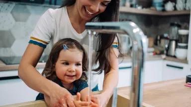 A mother helps her toddler wash their hands in the kitchen.