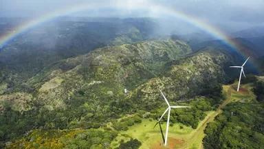 A rainbow arches over lush green mountains and wind turbines in a valley