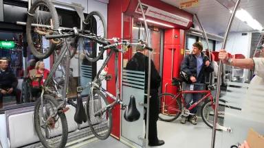 People ride in a passenger train car, while a man holds a bicylce and two more bicylces hang from a rack