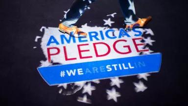 A person walks on the spot where a graphic is being projected onto the ground. The graphic is an outline of a U.S. map with the words "America's Pedge. #Weasrestillin".