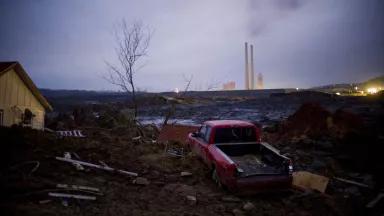 A yellow house and red pickup truck are among large pieces of debris partially buried in deep brown mud under dark clouds with two large industrial stacks visible in the distance