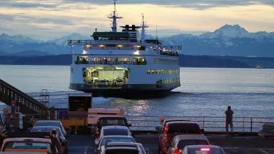 A large ferry carrying cars and passengers leaves a dock where more cars are lined up