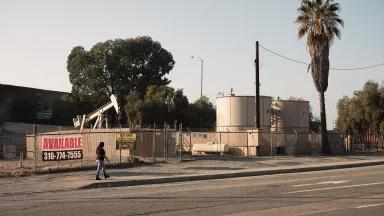 A woman walks on a city sidewalk past a fenced-in lot with an oil rig and two large holding tanks