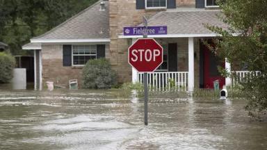 A stop sign stands above a flooded suburban street. In the background, a two-story home is flooded up to its front door and a water line on the side of the building reaches the first-story windows.