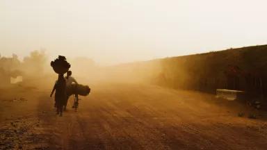 A man and a woman walk away from camera down a big, expansive, dusty road. The man pushes a bicycle with goods slung over the seat, and the woman carries a basket on her head.